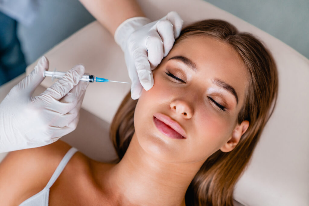 A woman with closed eyes receiving a cosmetic injection in her forehead. She is lying down and wearing a white tank top. A gloved hand is administering the injection with a syringe. Her hair is pulled back, and she appears relaxed. The setting is clinical and serene.